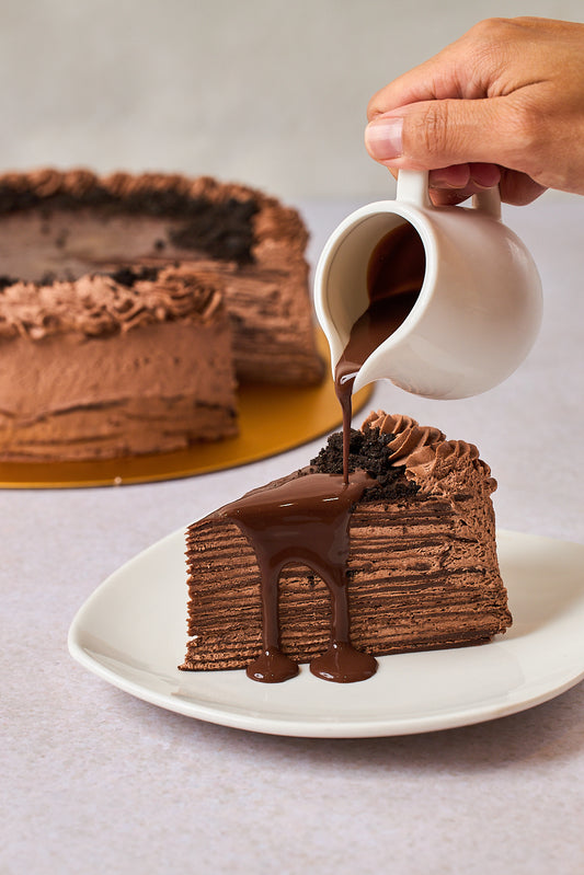 a hand pouring choco sauce on a piece of chocolate crepe cake